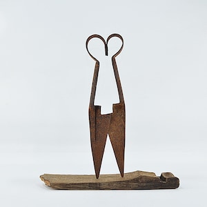 Könnte beinhalten: Rostige Metallschere mit herzförmigen Griffen, auf einem Stück verwittertem Holz stehend. Die Schere hat ein langes, schlankes Design, möglicherweise aus der Zeit. Die Gesamtästhetik ist rustikal und industriell.