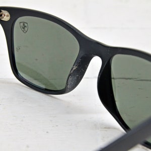 K&ouml;nnte beinhalten: Schwarze Sonnenbrille mit gr&uuml;nen Gl&auml;sern und einem silbernen Ferrari-Logo auf dem rechten Glas. Die Sonnenbrille ist aus Kunststoff und hat einen klassischen Stil.