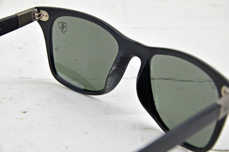 K&ouml;nnte beinhalten: Schwarze Sonnenbrille mit gr&uuml;nen Gl&auml;sern und einem silbernen Ferrari-Logo auf dem rechten Glas. Die Sonnenbrille ist aus Kunststoff und hat einen klassischen Stil.