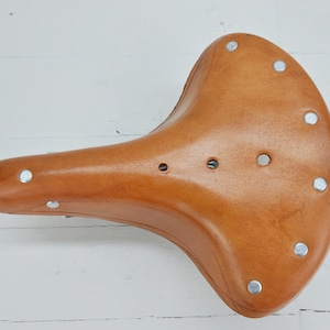 May include: A light brown leather bicycle saddle with a curved design. The saddle has a series of silver rivets around the edges and three small holes near the center. The saddle is designed for comfort and durability.