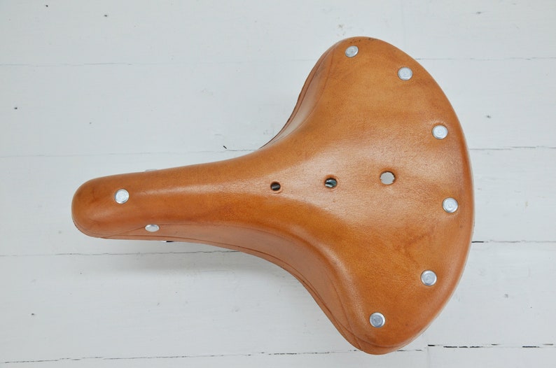 May include: A light brown leather bicycle saddle with a curved design. The saddle has a series of silver rivets around the edges and three small holes near the center. The saddle is designed for comfort and durability.