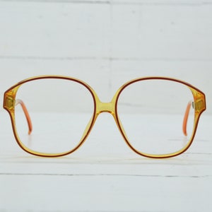 Op de afbeelding: Een paar amberkleurige brillen met een rood montuur. De bril heeft een ronde, oversized vorm.