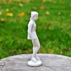 以下が含まれることがあります： 木製の台座の上に置かれた白い磁器製のフィギュアスケーターの置物。フィギュアは片足を伸ばしたスケートのポーズをとっている。