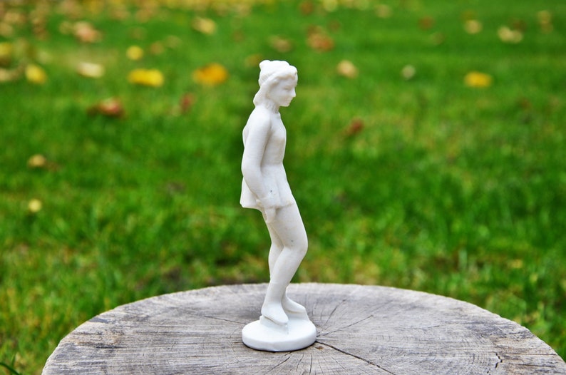 以下が含まれることがあります： 木製の台座の上に置かれた白い磁器製のフィギュアスケーターの置物。フィギュアは片足を伸ばしたスケートのポーズをとっている。