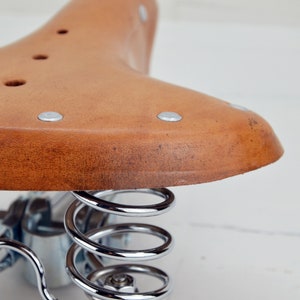 May include: Close-up of a vintage bicycle saddle. The saddle is made of brown leather with silver rivets and a distressed appearance. It features a curved shape and is supported by shiny, chrome-plated springs.