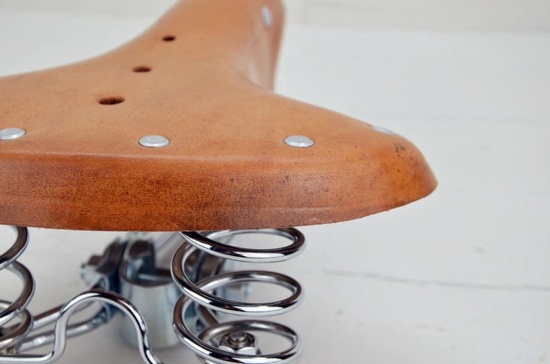 May include: Close-up of a vintage bicycle saddle. The saddle is made of brown leather with silver rivets and a distressed appearance. It features a curved shape and is supported by shiny, chrome-plated springs.