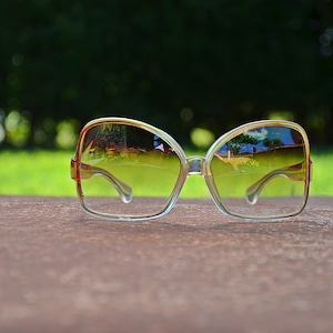 May include: A pair of large, clear, plastic framed sunglasses with brown lenses. The sunglasses are reflecting a blurry image of trees and a building.