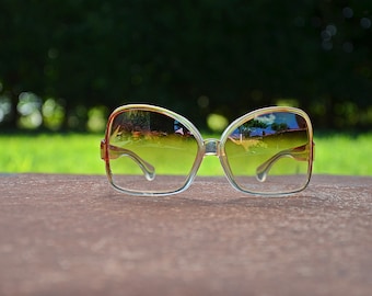 May include: A pair of large, clear, plastic framed sunglasses with brown lenses. The sunglasses are reflecting a blurry image of trees and a building.