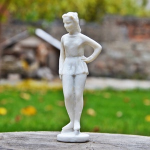 以下が含まれることがあります： 白い磁器製の女性像。女性はドレスを着ており、台座の上に立っている。女性は片手を腰に、もう片方の手を横に添えてポーズをとっている。