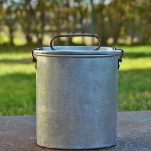 Peut inclure: Une casserole de camping en métal argenté avec une poignée en métal noir. La casserole est posée sur une surface grise avec un arrière-plan vert flou.