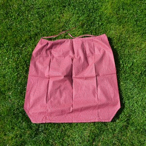 Puede incluir: Un delantal rojo con un estampado de lunares blancos, sobre hierba verde. El delantal tiene forma rectangular con una cinta en la parte superior. La tela está doblada y arrugada.