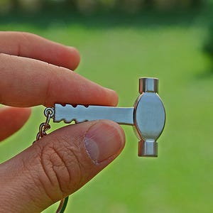 Op de afbeelding: Een kleine, zilverkleurige metalen sleutelhanger in de vorm van een hamer. Het handvat heeft een reeks inkepingen langs de bovenrand, terwijl de kop afgerond en glad is. Een kleine metalen ring is aan het uiteinde van het handvat bevestigd.