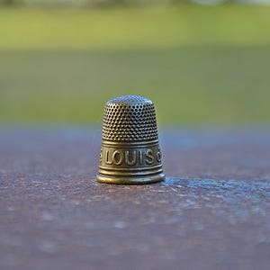 Peut inclure: Un dé à coudre en métal doré avec le mot "LOUIS" gravé sur le côté.