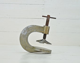 May include: A silver metal C-clamp with a rusty screw and a handle. The clamp is open and ready to be used.