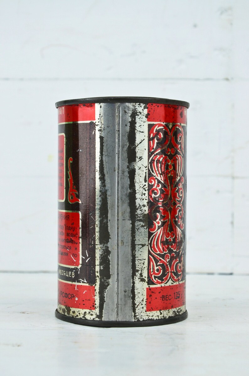 May include: A vintage metal tin with red and black lettering and a decorative design. The tin is labeled "Вес 125" and has text in Russian.