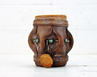 May include: A brown leather dice cup with green gemstone accents. The cup has a round, wooden lid.