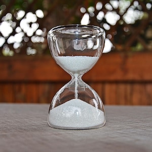May include: A clear glass hourglass with white sand, sitting on a grey surface.
