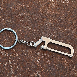 May include: A silver-toned metal keychain featuring a miniature hacksaw charm. The charm is attached to a standard circular split ring by a short metal chain. The hacksaw design includes a handle, a frame, and a serrated blade in a metallic finish.