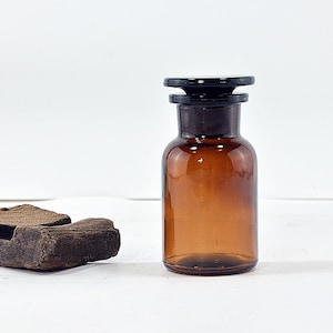 Puede incluir: Una botella de boticario vintage de vidrio ámbar con un tapón negro. La botella tiene un cuerpo redondeado y un cuello estrecho. Un trozo de madera envejecida está en el fondo. La botella probablemente se usa para almacenar líquidos o polvos.