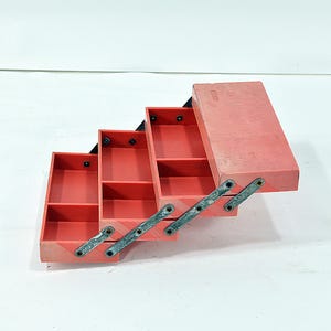 May include: A red wooden toolbox with multiple compartments, open and angled. The toolbox has metal hinges and a black interior. The box is empty and sits on a white surface. The toolbox is designed for storing tools and small items.