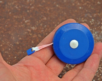 Puede incluir: Una cinta métrica retráctil azul con un botón blanco y una cinta métrica rosa y blanca que se extiende desde la caja. La cinta métrica se sostiene en una mano.