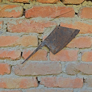 May include: A rusty, vintage cleaver with a long, pointed handle is mounted on a weathered brick wall. The cleaver's blade is rectangular with a small hole near the top. The bricks are reddish-orange with gray mortar.
