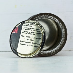 May include: A round metal tin with a black and white label. The label reads "залить горячек размешать. Сахар, молоко илк ки добавить по вкусу. Банку с кофе хранкт плотно закрытой. ГОСТ 5.863-71 Цена 2 руб. Вес 100 г. Срок хранения в месяцев. Хранить в сухом месте."