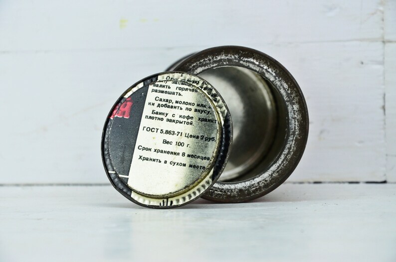 May include: A round metal tin with a black and white label. The label reads "залить горячек размешать. Сахар, молоко илк ки добавить по вкусу. Банку с кофе хранкт плотно закрытой. ГОСТ 5.863-71 Цена 2 руб. Вес 100 г. Срок хранения в месяцев. Хранить в сухом месте."