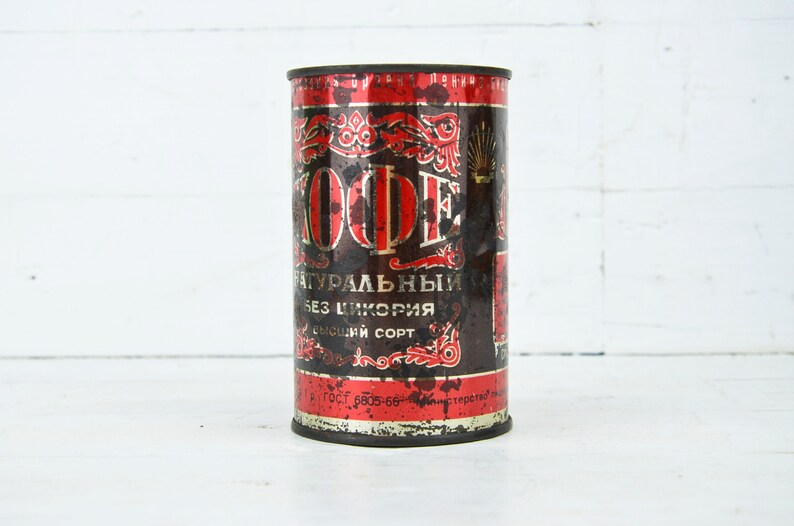 May include: A vintage red and black metal can with the word "KOФE" in gold lettering. The can is labeled "натуральный без цикория высший сорт" and has a faded, worn look.