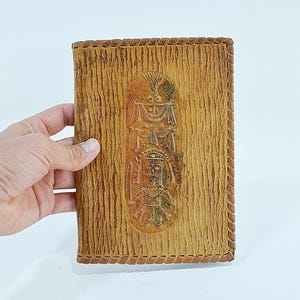 May include: A tan leather notebook cover with a textured, bark-like surface and a central oval embossed design featuring a stylized figure and decorative elements. The edges are finished with a braided leather lacing detail along the perimeter.