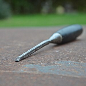 May include: A metal chisel with a black handle, lying on a weathered, rusty surface. The chisel's blade is angled and appears sharp. The background is blurred, with hints of green foliage.
