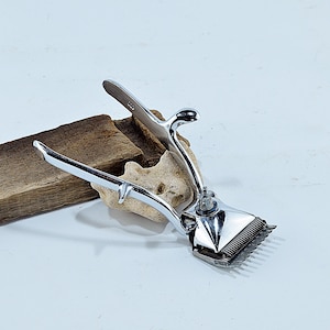 Könnte beinhalten: Ein verchromter Vintage-Haarschneider, der auf einem Stück verwittertem Holz liegt. Der Haarschneider verfügt über einen Hebel, einen Griff und Schneidklingen. Der Hintergrund ist eine schlichte weiße Oberfläche.