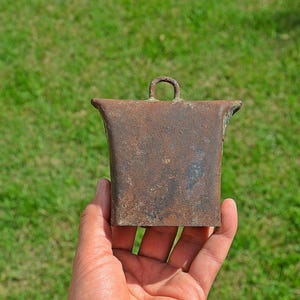 May include: A rusty metal cowbell with a rounded top and a handle. The bell is brown and has a worn, aged appearance.