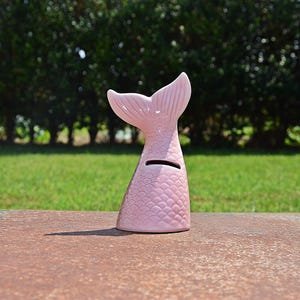 May include: Pink ceramic mermaid tail piggy bank with a coin slot on the top. The tail has a scaly texture.