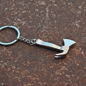 Op de afbeelding: Een zilverkleurige metalen sleutelhanger met een miniatuur bijlvormige bedel die met een kort kettinkje aan een ronde splitring is bevestigd. De bijlbedel heeft een gladde, gepolijste afwerking met een gebogen snijvlak en een steel.
