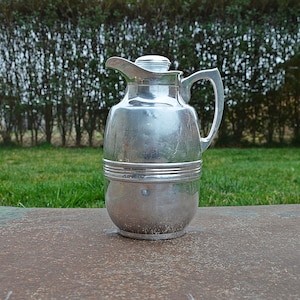 Può includere: Un thermos d'argento con un coperchio e un manico. Il thermos è appoggiato su una superficie di metallo arrugginito.
