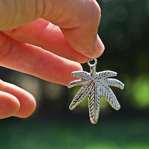 Puede incluir: Un amuleto de plata con forma de hoja de marihuana. El amuleto está siendo sostenido por una mano sobre un fondo verde.