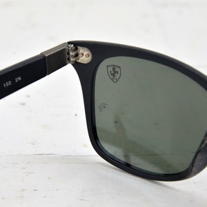 K&ouml;nnte beinhalten: Schwarze Sonnenbrille mit gr&uuml;nen Gl&auml;sern und einem Ferrari-Logo auf der rechten Linse. Die Sonnenbrille hat einen schwarzen Rahmen und ein schwarzes Band.