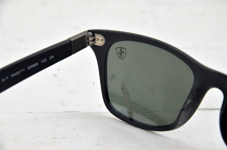 K&ouml;nnte beinhalten: Schwarze Sonnenbrille mit gr&uuml;nen Gl&auml;sern und einem Ferrari-Logo auf der rechten Linse. Die Sonnenbrille hat einen schwarzen Rahmen und ein schwarzes Band.