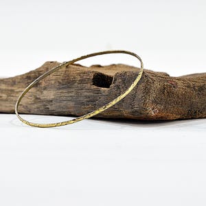 May include: A thin, gold-colored bangle bracelet with a textured surface rests on a piece of weathered wood. The bracelet is circular and appears to be made of metal. The wood has a rough, natural appearance.