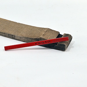 May include: A red pencil with the word "FESTA" printed on it rests on a dark brown wooden block. The block has a rough, textured surface and a rectangular shape. The pencil is positioned diagonally.