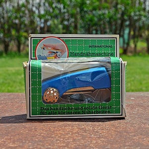 Op de afbeelding: Een vintage blauwe handnaaimachine in de originele doos. De doos is groen en wit met de woorden "INTERNATIONAL Handnahmaschine" en Duitse tekst. De naaimachine heeft een gouden wiel en is zichtbaar door een helder plastic venster.