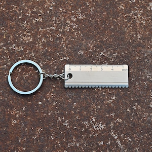 May include: A silver-colored metal keychain featuring a circular split ring attached by a short chain to a rectangular tool. The tool has a straight edge with markings from 0 to 5 cm and a serrated edge along the bottom side of the metal plate.