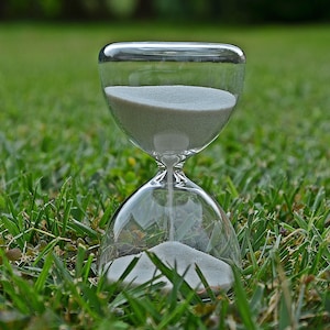May include: A clear glass hourglass with white sand, sitting on a bed of green grass.