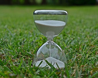 May include: A clear glass hourglass with white sand, sitting on a bed of green grass.