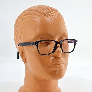 May include: A pair of black-framed spectacles with rectangular lenses is displayed on a mannequin head. The glasses have a classic design with a dark finish. The mannequin head is a light brown colour.