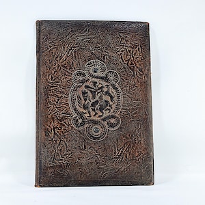 May include: A vintage, brown leather-bound book with an embossed design. The cover features a central medallion with a detailed relief of figures, surrounded by ornate scrollwork. The leather has a textured, aged appearance.