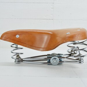 May include: A brown leather bicycle seat with a chrome spring suspension system. The seat is a classic design, with a curved shape and rivets. The springs are coiled and provide cushioning. The seat is designed for comfort and style.