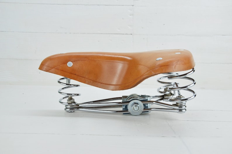 May include: A brown leather bicycle seat with a chrome spring suspension system. The seat is a classic design, with a curved shape and rivets. The springs are coiled and provide cushioning. The seat is designed for comfort and style.