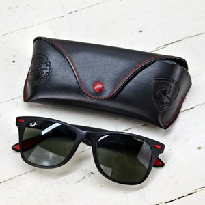 K&ouml;nnte beinhalten: Schwarze Sonnenbrille mit roten Akzenten und einem schwarzen Lederetui mit roten N&auml;hten. Das Etui hat ein gepr&auml;gtes Ferrari-Logo.
